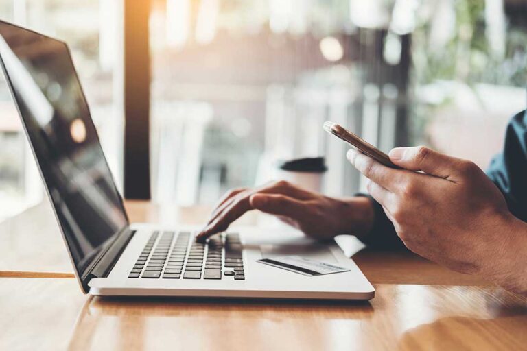 Person reviewing monthly expenses at a kitchen table with a notebook, calculator, receipts, and laptop.